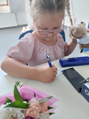 Dziewczynka siedzi przy stoliku i rysuje na kartce, na stole leżą kwiatki