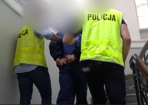 policjanci w kamizelkach odblaskowych prowadzący zatrzymanego mężczyznę po schodach