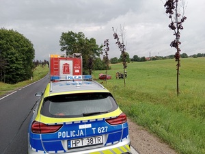 radiowóz policyjny stojący na drodze, przed nim wóz strażacki, w rowie po prawej stronie stojący czerwony samochód osobowy