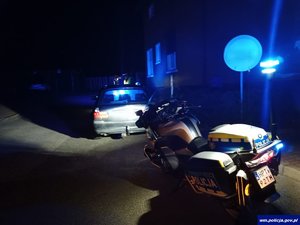 zdjęcie zrobione nocą. Motocykl policyjny oświetla samochód osobowy stojący przed nim