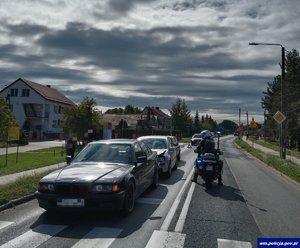 stojące na drodze przed przejściem dla pieszych trzy samochody osobowe jeden z drugim. Po ich prawej stronie jedzie motocykl policyjny z włączonymi sygnałami świetlnymi