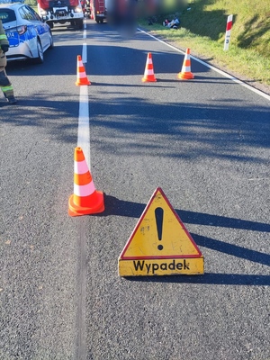 znak drogowy "wypadek" stojący na drodze, dalej z lewej strony na jezdni radiowóz policyjny, przed nim stoi wóz strażacki, po prawej stronie w rowie leży motocykl, miejsce zdarzenia drogowego odgrodzone jest pomarańczowymi pachołkami