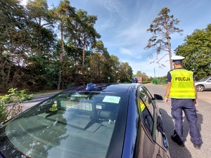 z przodu na parkingu stoi nieoznakowany radiowóz z napisem wyświetlanym Policja, przy nim stoi policjant ruchu drogowego