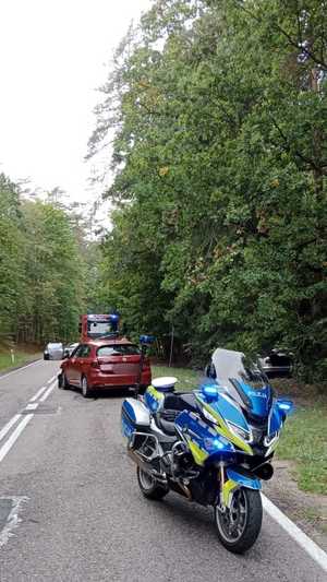 stojący na drodze motocykl policyjny, za nim stoi czerwony samochód osobowy a jeszcze dalej wóz strażacki. W rowie po prawej stronie widać samochód osobowy koloru srebrnego