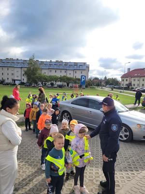 na parkingu stoją dzieci w rzędzie w parach, na ramieniu pierwszego dziecka policjant trzyma dłoń, z lewej strony stoi kobieta