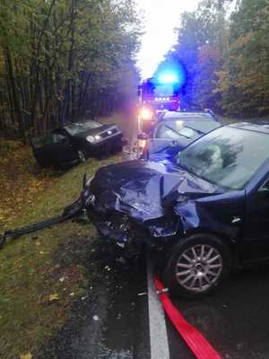 rozbite auto stojące na drodze, kolejne auto z rozbitym przodem stoi w rowie, za nimi stoii straż pożarna