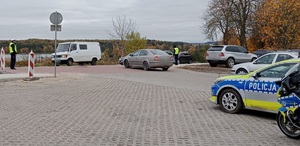 na parkingu przy cmentarzu z prawej strony stoi motocykl policyjny, obok niego radiowóz, z przodu stoją policjanci którzy kierują ruchem drogowym