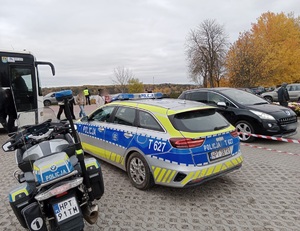 motocykl policyjny, radiowóz policyjny stojące na parkingu, przed nimi stoi autobus a przy nim ludzie