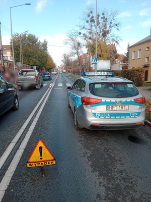 zdjęcie stojącego na drodze radiowozu, z lewej strony przejeżdżające pojazdy a za radiowozem znak drogowy z napisem wypadek