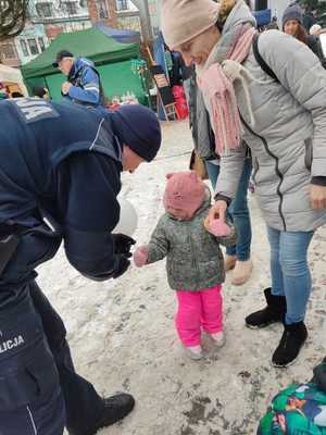 Policjant pochyla się nad dzieckiem, kobieta trzyma dziecko za rękę. Osoby znajdują się na zewnątrz