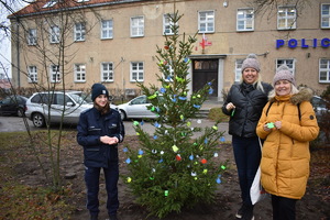 przed budynkiem Komendy Powiatowej Policji w Mrągowie z lewej strony stoi policjantka obok niej wkopana jest choinka na której zawieszone są odblaski. Obok choinki z prawej strony stoją dwie kobiety, które trzymają w ręku odblaski