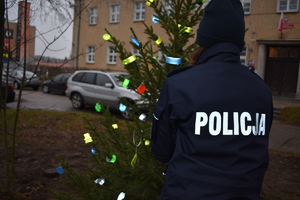 przed budynkiem komendy policjantka stoi tyłem, na kurtce ma napis policja. stoi przed choinką na której są odblaski