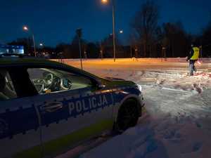 wieczorową porą przy drodze na śniegu stoi radiowóz, przed nim stoi policjant