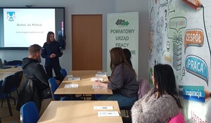 na sali stoi policjantka przed słuchaczami w tle wyświetlana jest prezentacja z hasłem Dobór do Policji