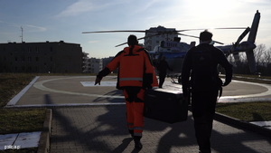 idący w kierunku lądowiska helikoptera i stojącego tam helikoptera i ratownik medyczny z drugim mężczyzną niosący skrzynię w kierunku helikoptera