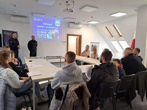 na sali siedzą słuchacze, a przed nimi stoi 2 policjantów, na ścianie wyświetlana jest prezentacja