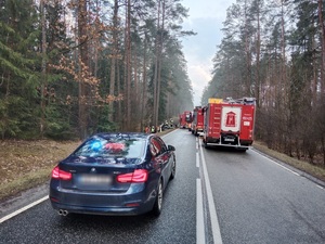na drodze z lewej strony stoi nieoznakowany radiowóz, na prawym pasie ruchu stoi straż pożarna, w dali widać miejsce zdarzenia drogowego