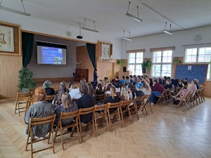 maturzyści siedzący na auli, przed nimi policjantka która prezentuje wyświetlaną prezentację