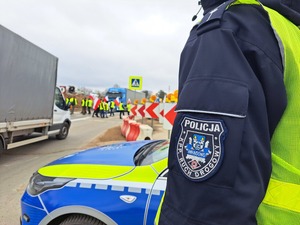 na pierwszym planie stoi radiowóz, obok niego bokiem stoi policjant i na mundurze widać napis Komenda Powiatowa Policji w Mrągowie. W tle widać ciągniki i protestujących rolników