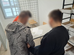 w pomieszczeniu dla osób zatrzymanych stoi trzech mężczyzn, jeden z nich ma założone kajdanki na ręce, a drugi go trzyma