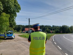 policjant ruchu drogowego stojący przy drodze z miernikiem prędkości, z lewej strony stoi radiowóz