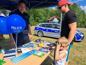 policjant stoi przy stoisku profilaktycznym a przed nim stoją uczestnicy pikniku