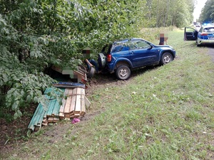 stojący w rowie samochód z przyczepką z której spadły deski, po prawej stronie na drodze stoi radiowóz