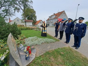 policjanci i ksiądz stoją przy obelisku Marka Cekały. Policjanci oddają honory