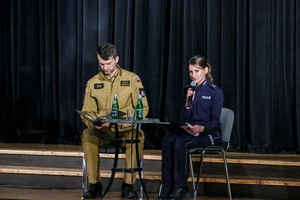 siedząca na scenie przy stoliku policjantka a obok niej strażak. Policjantka czyta do mikrofonu bajkę
