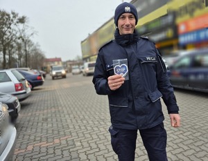 policjant trzymający w ręku serduszko- zawieszkę do samochodu z napisami przypominającymi kierującym o zachowaniu ostrożności przy zbliżaniu się do przejść dla pieszych