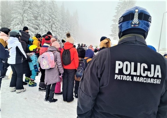 zdjęcie na którym tyłem stoi policjant który ma napis na plecach Policja Patrol Narciarski a w tle widać osoby na nartach