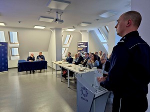 Komendant stoi przy mównicy a w tle widać osoby biorące udział w odprawie