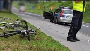 leżący na poboczu rower a w tle radiowóz stojący na drodze i stojący na chodniku policjant