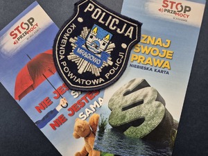 emblemat Komendy Powiatowej Policji w Mrągowie a w tle ulotki dotyczące przemocy domowej