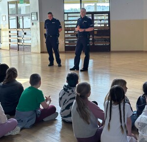 uczniowie siedzący na hali i policjanco stojący przed nimi