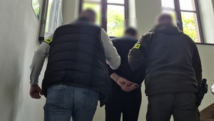 dwóch nieumundurowanych policjantów, którzy prowadzą zatrzymanego mężczyznę po schodach w górę. Mężczyzna ma założone kajdanki na ręce trzymane z tyłu