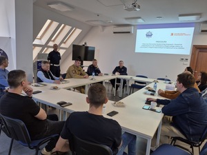 osoby siedzące przy stole oraz stojący przy nich policjant. Na ścianie wyświetlana prezentacja