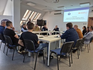 osoby siedzące przy stole oraz stojący przy nich policjant. Na ścianie wyświetlana prezentacja