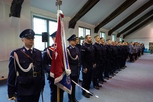 policjanci w mundurach galowych na sali. Jeden z policjantów trzyma sztandar Komendy Wojewódzkiej Policji w Olsztynie