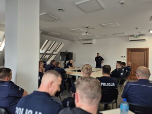 siedzący na sali policjanci i stojący przed nimi Rzecznik Prasowy Komendanta Wojewódzkiego Policji w Olsztynie