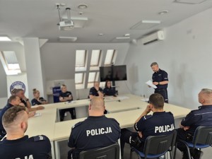 siedzący na sali policjanci i stojący przed nimi Rzecznik Prasowy Komendanta Wojewódzkiego Policji w Olsztynie