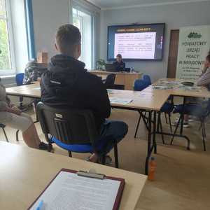 policjantka siedzi przed komputerem, za nią wyświetlana jest prezentacja a przed nią siedzą słuchacze