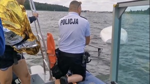 policyjny wodniak na policyjnej łódce za pomocą sznura ściąga łódź, która jest przewrócona w jeziorze do góry dnem