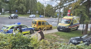 przy ulicy stoi zaparkowana karetka, radiowóz oraz samochód. Wokół jest kilka osób i policjant