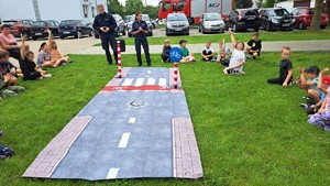 policjantka i policjant stoją przed rozłożonym przejściem dla pieszych a wokół nich na trawie siedzą dzieci