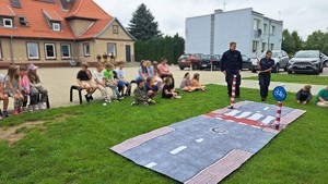 policjantka i policjant stoją przed rozłożonym przejściem dla pieszych a wokół nich na trawie siedzą dzieci