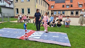 policjantka i dziewczynka stoją przed rozłożonym przejściem dla pieszych a wokół nich na trawie siedzą dzieci.