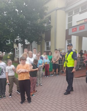 policjant stoi przed budynkiem szkoły, a ze szkoły wychodzą uczniowie