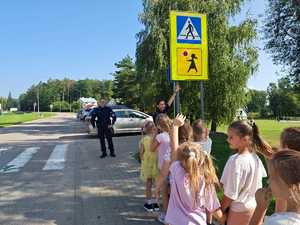 przy przejściu dla pieszych stoi policjant przed nim policjantka pokazuje dzieciom znak drogowy