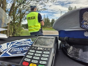leżące na podszybiu samochodu czapka policjanta ruchu drogowego, terminal płatniczy i naklejki z napisem &quot;Mistrzu zwolnij&quot; w tle przez szybę widać policjanta ruchu drogowego